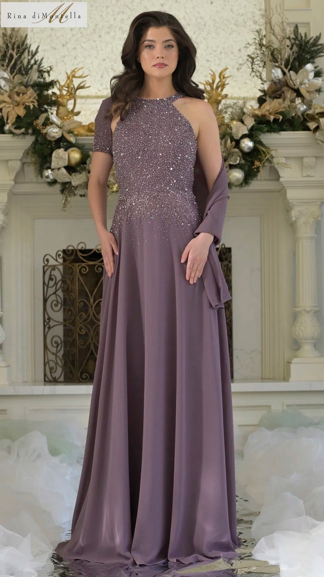 A woman stands indoors in front of a decorated mantle, wearing the Rina Di Montella RD3304 long formal dress in Victorian Lilac—a halter gown with sequin detailing and a chiffon A-line skirt.